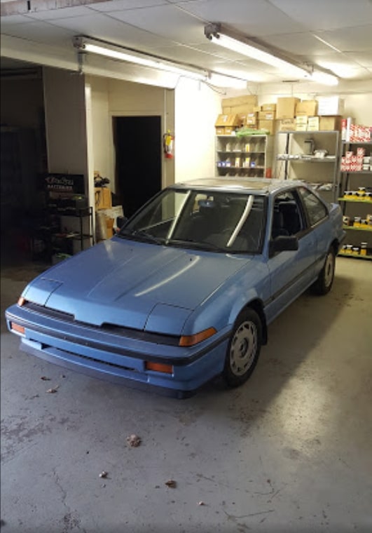 A blue car parked inside a garage, with spare parts arranged neatly on a rack in the background